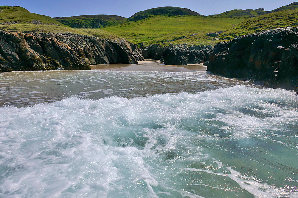 Picture of a wave breaking and rolling on to a beach between some rocks