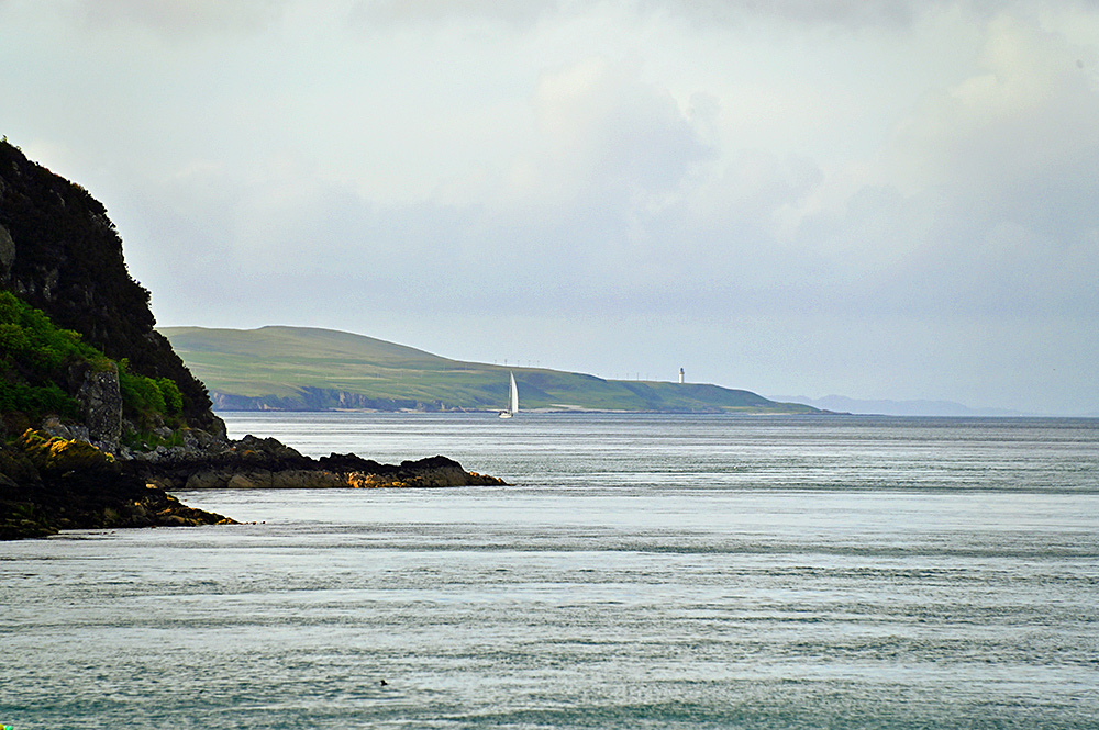 Picture of a sailing yacht off a shoreline with cliffs and bays, also a lighthouse visible on the horizon