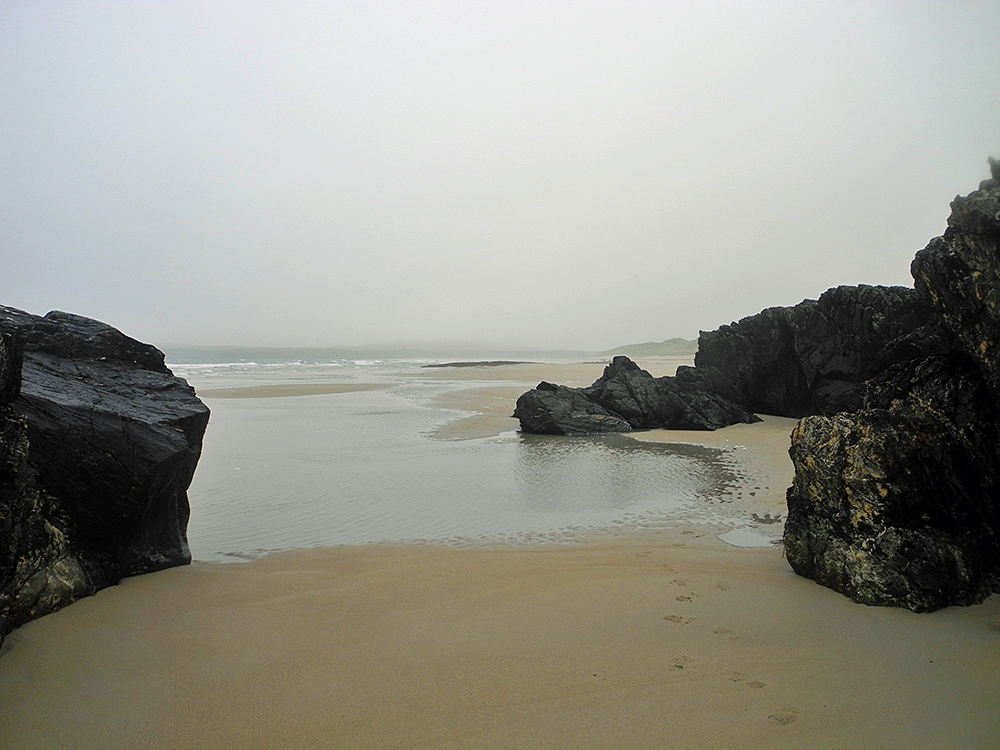 Picture of a view out from between some dark rocks over a sandy beach on a misty morning, the other side of the bay has disappeared in the mist