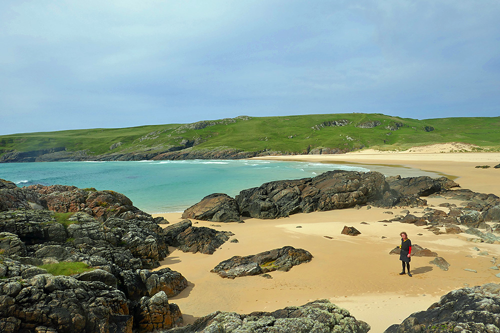 Picture of a view from low cliffs on to a bay with a beach and some rocks, a person standing on one side