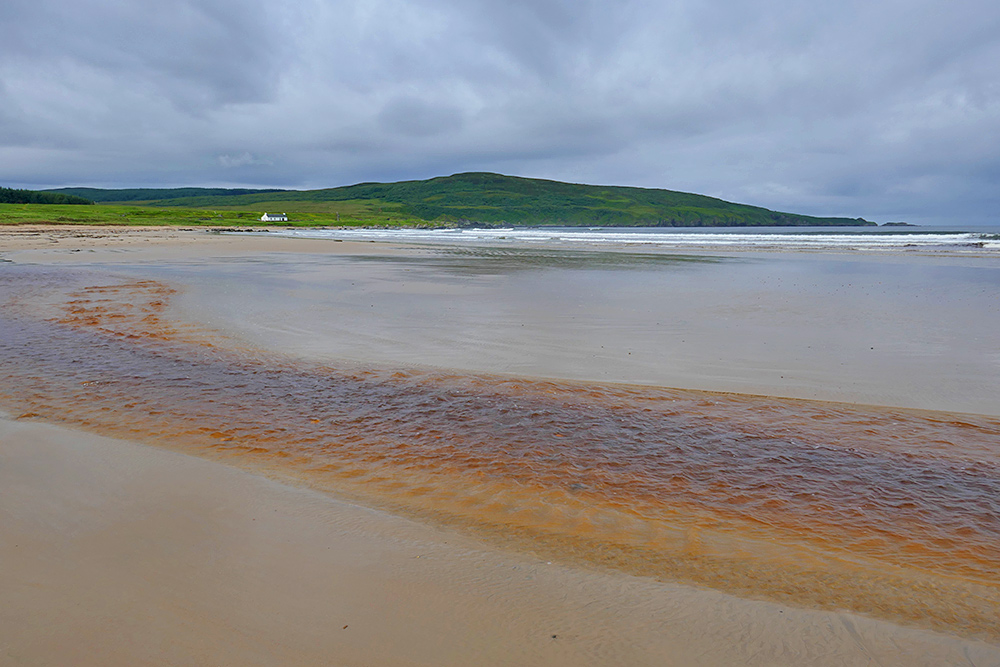 Picture of a small peaty river flowing over a sandy beach, a cottage and green hills in the background