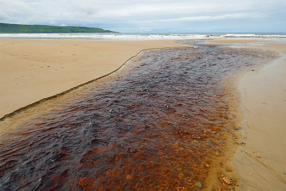 Picture of a small peaty river running over a sandy beach into the sea