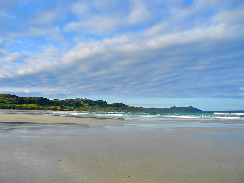 Picture of a wide sandy beach in some mild sunshine, some wavy clouds in the sky ahead