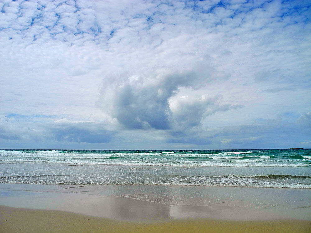 Picture of a view from a beach out to sea with waves rolling in, clouds above