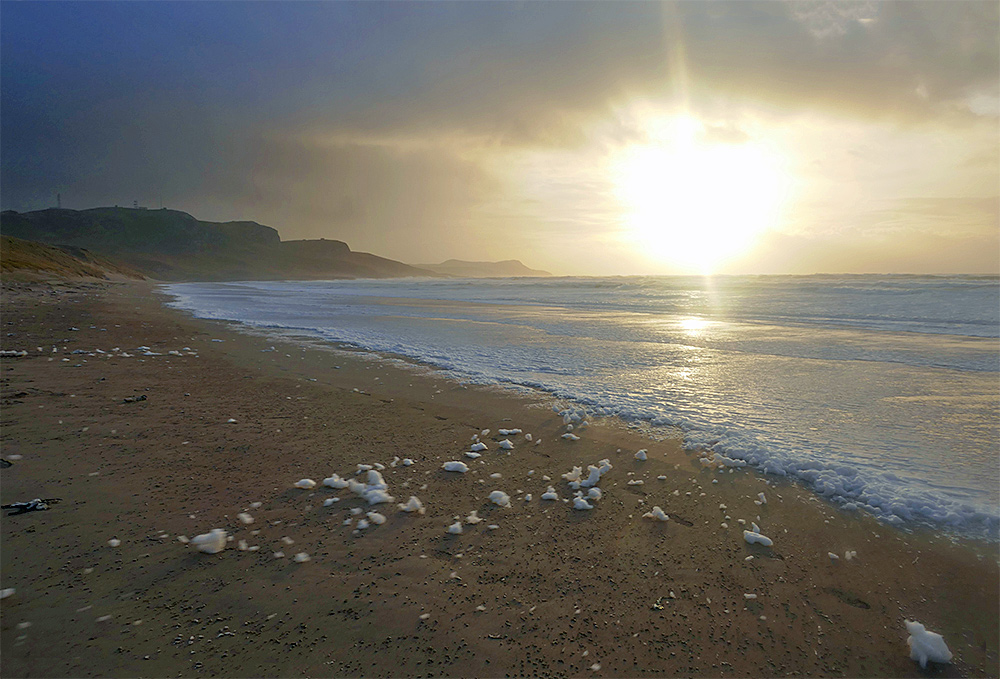 Picture of low afternoon sun over a bay with a beach on a stormy day, sea foam being blown across the beach