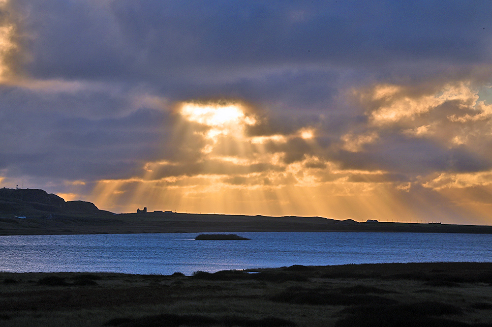 Picture of a sunburst through dark clouds over a rural landscape with a freshwater loch and scattered buildings including a church