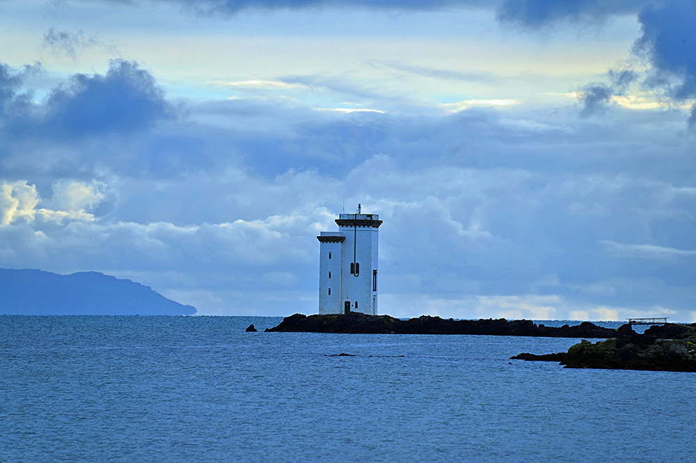 Picture of a square lighthouse on a rocky shore, the mull of a peninsula in the distance