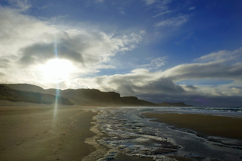 Picture of the sun breaking through dramatic clouds over crags next to a sandy beach
