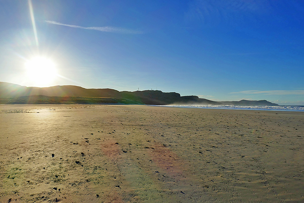 Picture of a low sun rising behind some crags shining over a sandy wide beach