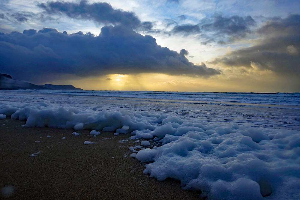 Picture of lots of sea foam on a long beach, some low afternoon sun behind a cloud