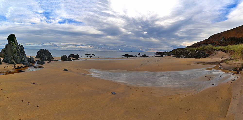 Panoramic picture of a sandy beach known as the Singing Sands with some rocks, land visible in the distance across the sea