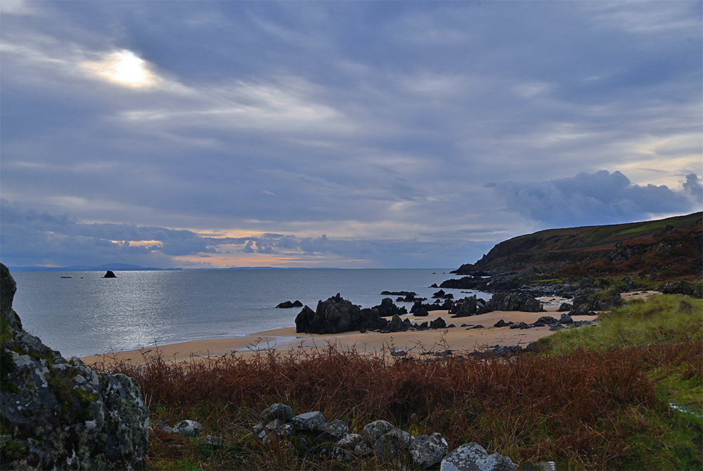 Picture of a view over a sandy beach interspersed with rocks, the sun breaking through the clouds. Another island visible on the horizon across the sea.
