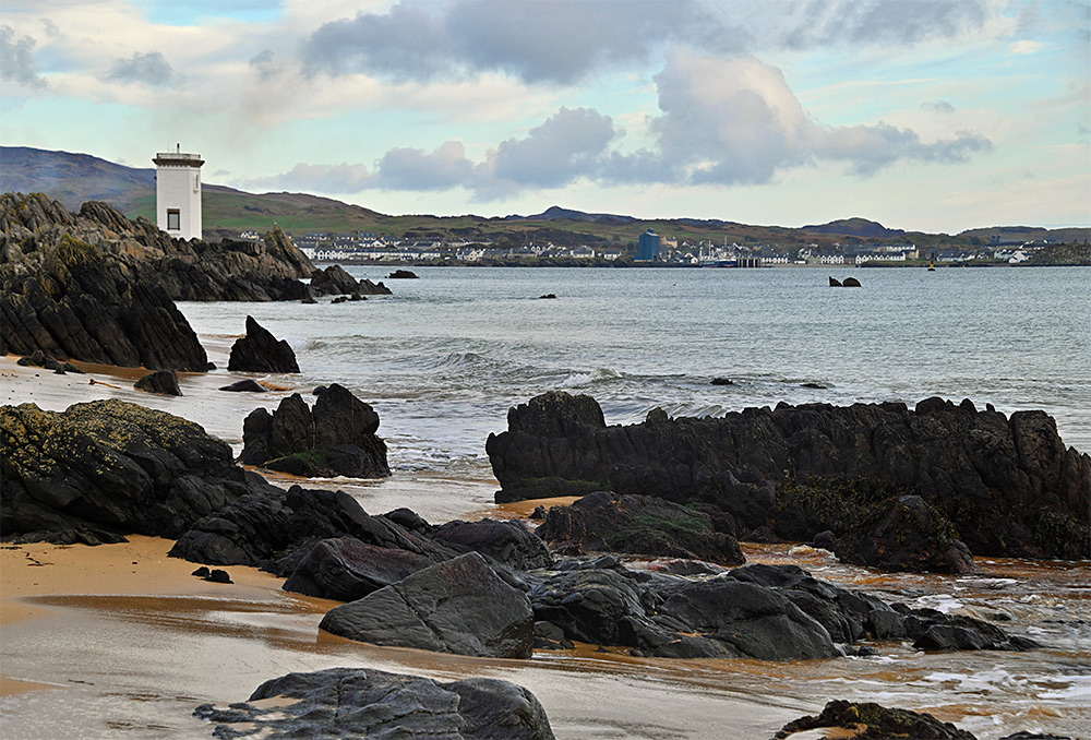 Picture of a view from a beach mixed with rocks to a square lighthouse and a coastal village across a wide bay