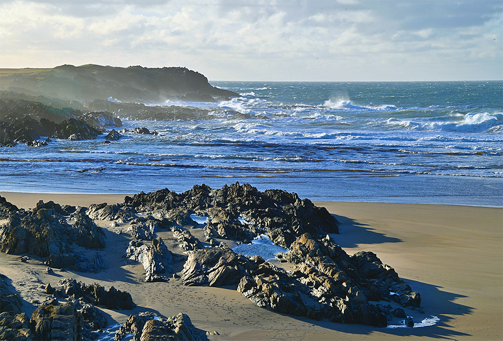 Picture of the end of a bay with cliffs, rocks and beach as well as waves rolling in