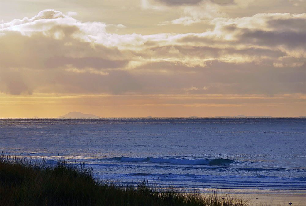 Picture of a view from a dune path over the sea to some distant hills just visible on the horizon. Colourful clouds in the November afternoon sky