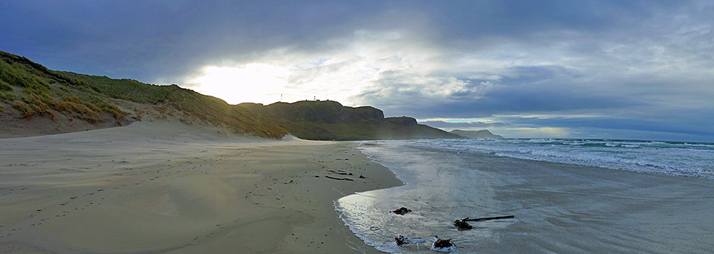 Panoramic picture of a wide sandy beach backed by dunes, dramatic clouds with the sun breaking through above