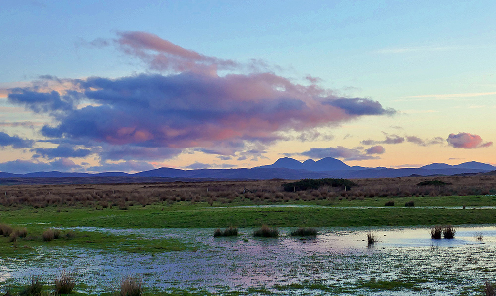 Picture of some colourful clouds in the November morning light over a rugged landscape behind a flooded field. Three round mountains in the distance.