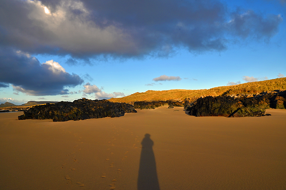 Picture of a sandy beach with some large rocky outcrops, sand dunes behind. Some dramatic dark clouds above