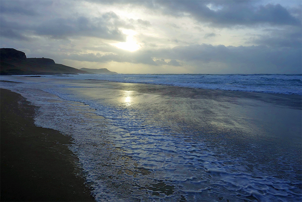 Picture of a wide beach on a stormy November afternoon, the wind has pushed water and foam high up the beach. The ripples of a guest can be seen on some shallow water