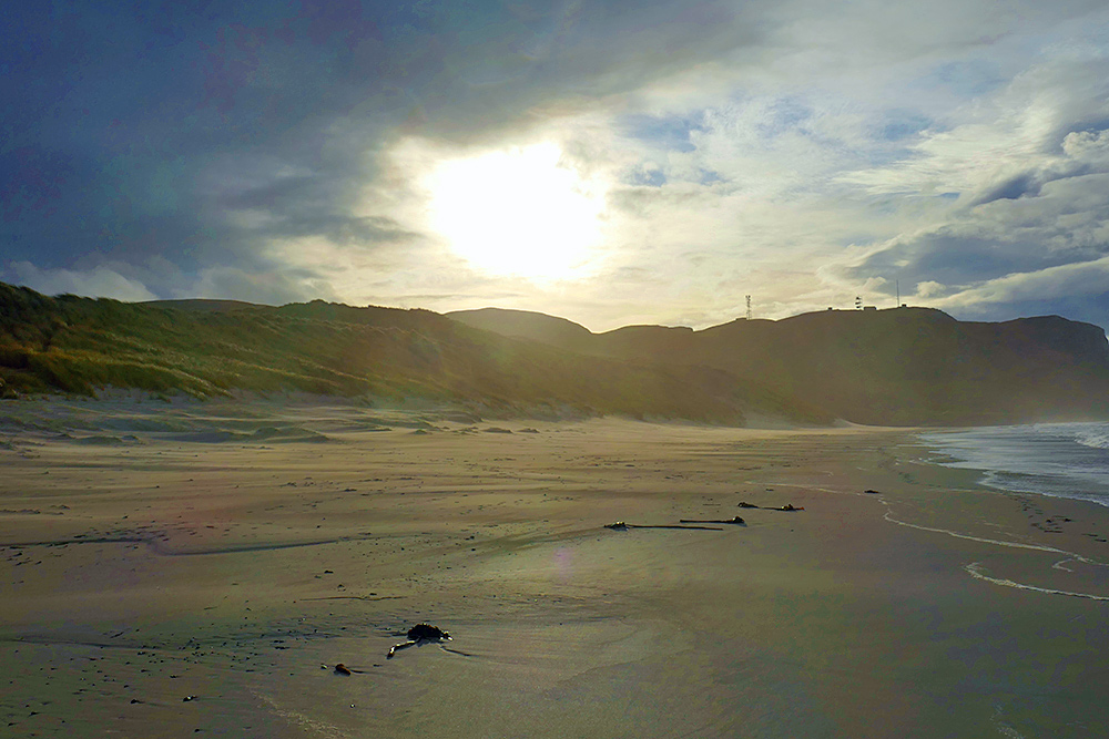 Picture of the sun breaking through clouds over dunes and crags behind a beach