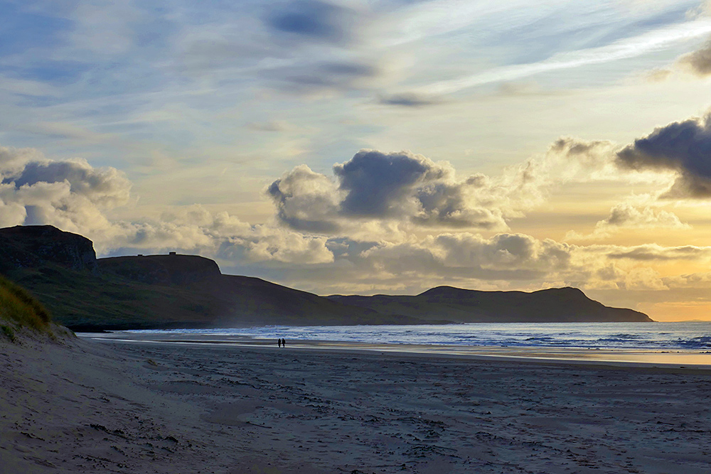Picture of two walkers on a sandy beach in a wide bay in the mild November late afternoon light not long before sunset