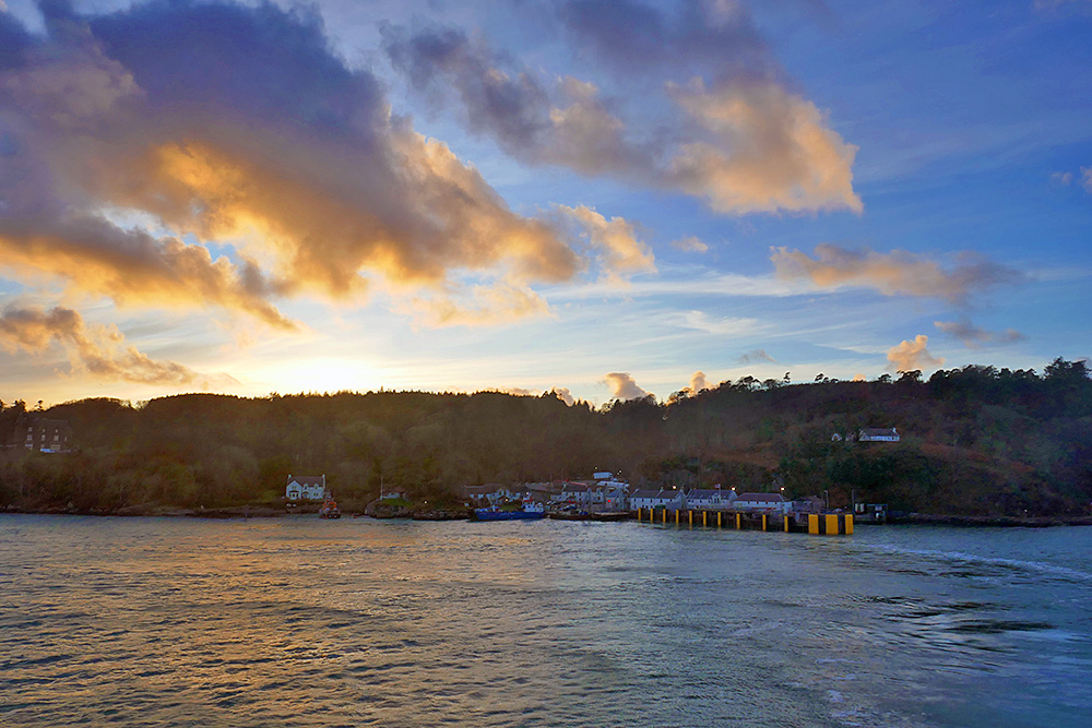 Picture of a tiny coastal village with a ferry terminal seen from the water under some colourful clouds