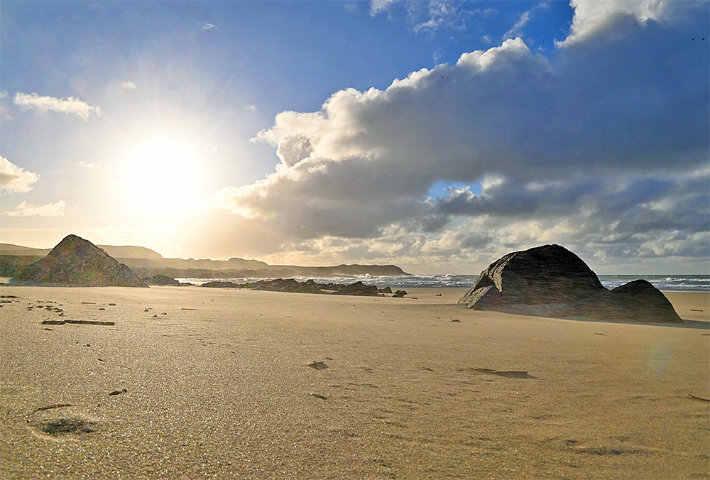 Picture of a low view over a sandy beach with some smooth rocks embedded in the sand, the low sun in the sky next to a band of clouds