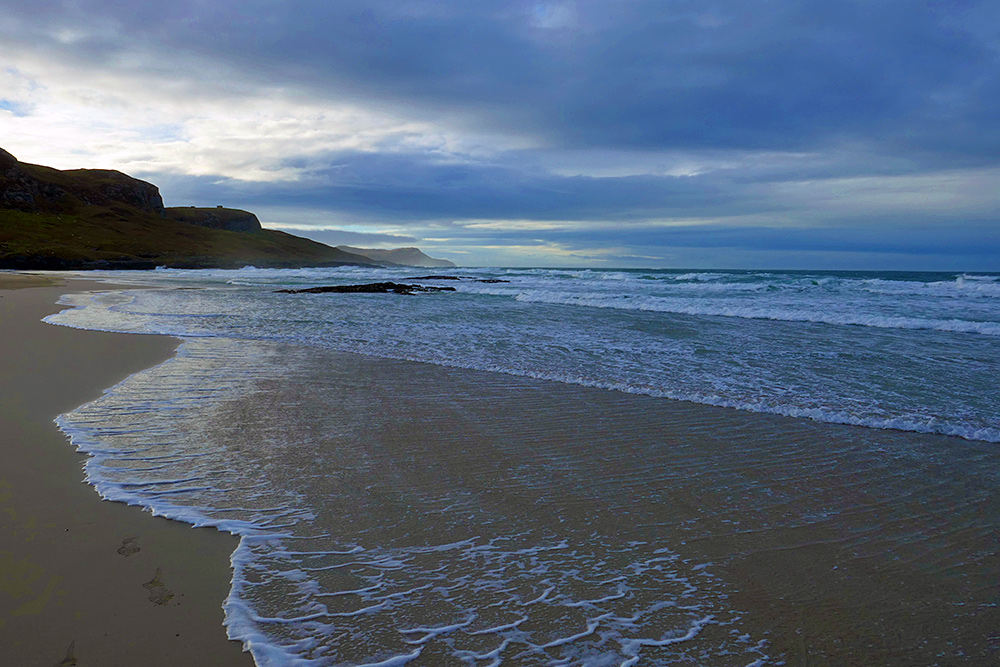 Picture of waves running out on a sandy beach with a rocky outcrop, dark clouds above