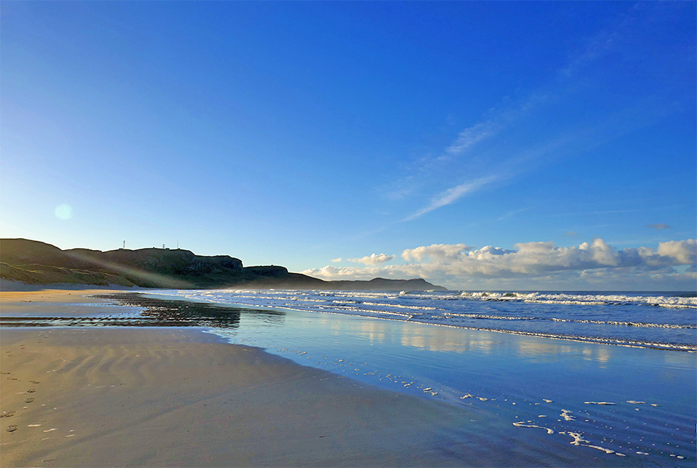 Picture of a big wide blue sky over a bay with a wide sandy beach and waves breaking as they roll in