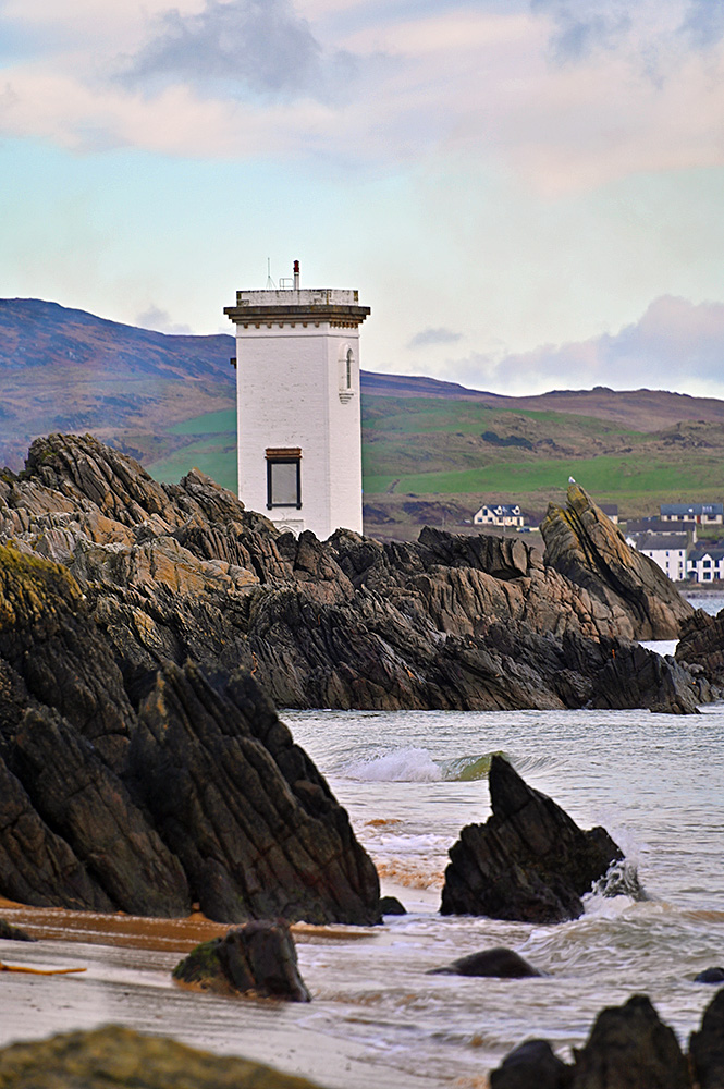 Picture of a square white lighthouse behind some rugged cliffs next to a sandy beach