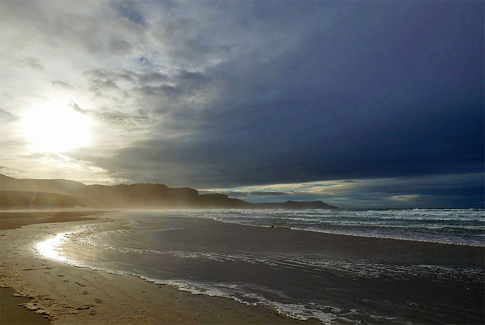 Picture of a wide sandy beach in a wide bay under dark clouds with a bright sun breaking through the clouds