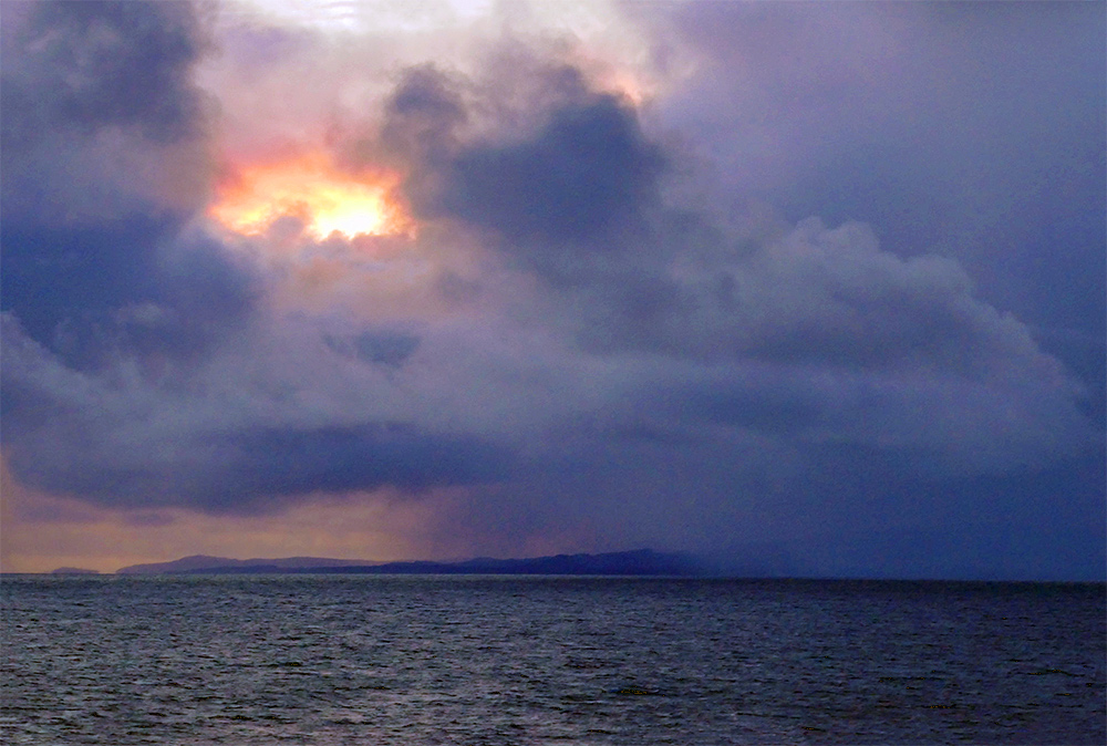 Picture of a view from a ferry to an island, the sun breaking through clouds on the left with there is heavy rain on the right