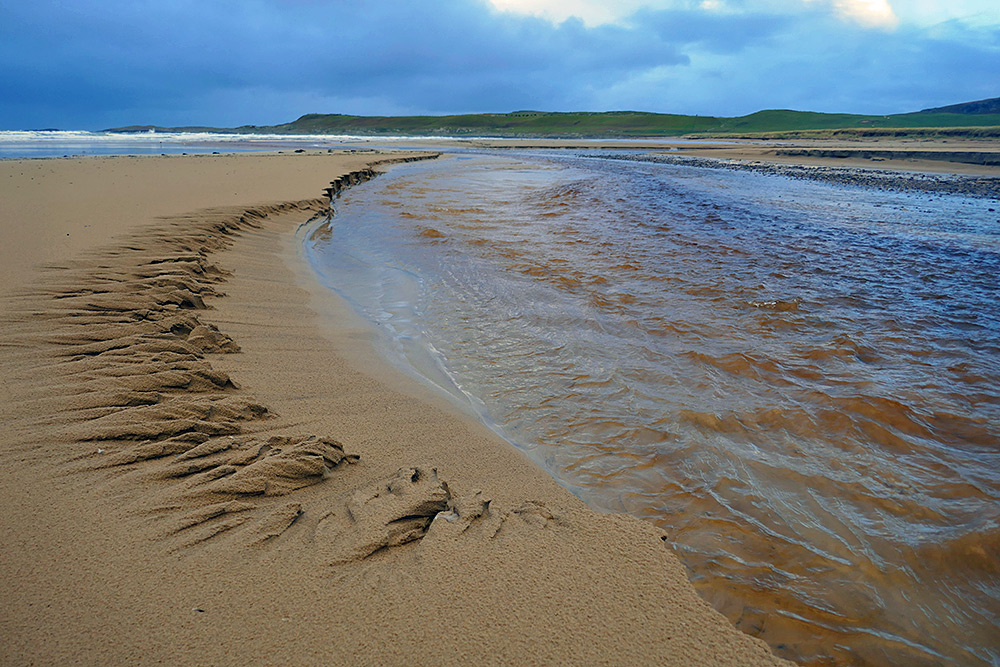 Picture of a burn (stream) flowing over a beach, erosion visible on the bank
