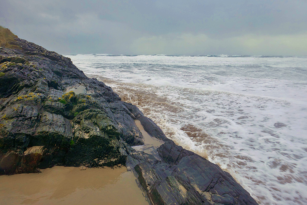 Picture of a storm wave rushing up a beach next to a rocky hillside
