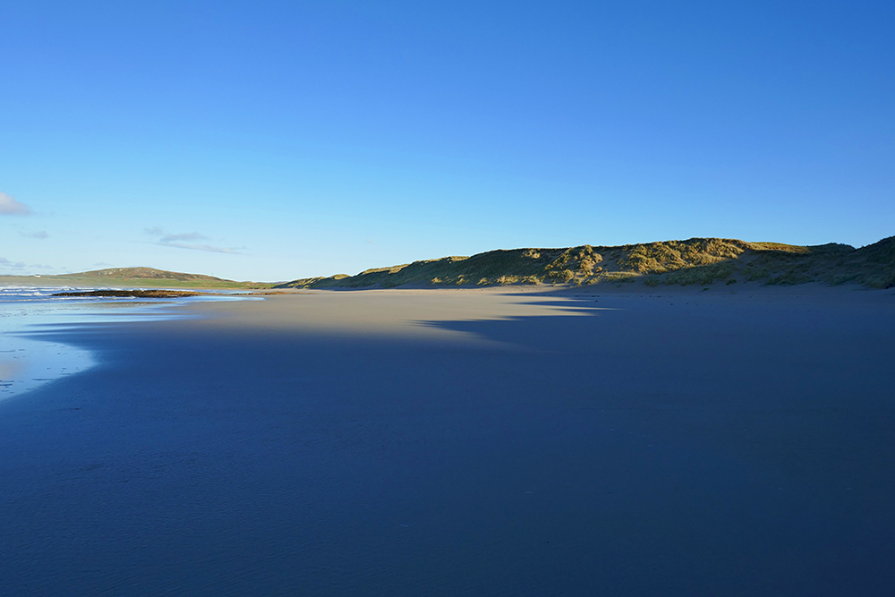 Picture of a view over a long wide sandy beach from out of the shadows of crags behind the viewer