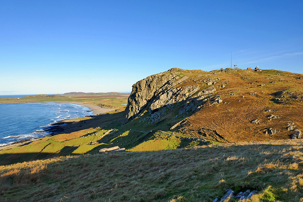 Picture of a sandy beach below some steep crags with some buildings (an old radar station now used for telecoms) on the top on a sunny November day