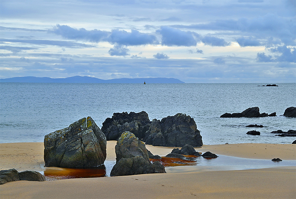 Picture of a view from a beach with some rocks to a distant peninsula, also a lone buoy in the distance