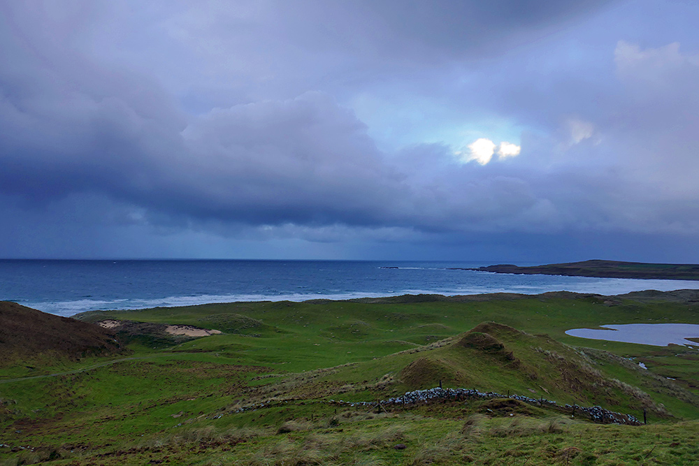 Picture of a view over dunes out to a wide bay under heavy clouds, rain in the distance