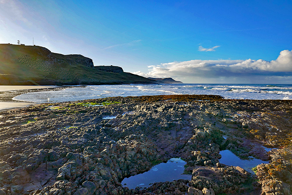 Picture of a view from a rocky outcrop in a beach over the end of a bay with a sandy beach and a rugged shoreline stretching out into the distance