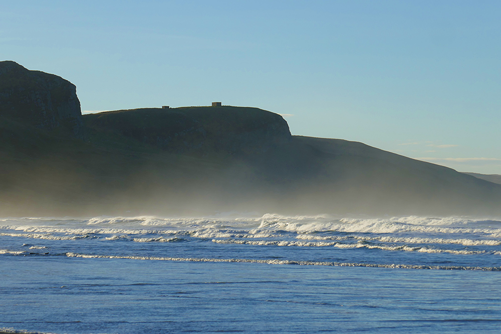 Picture of waves with a lot of spray and mist in the air above them, dark crags in the background