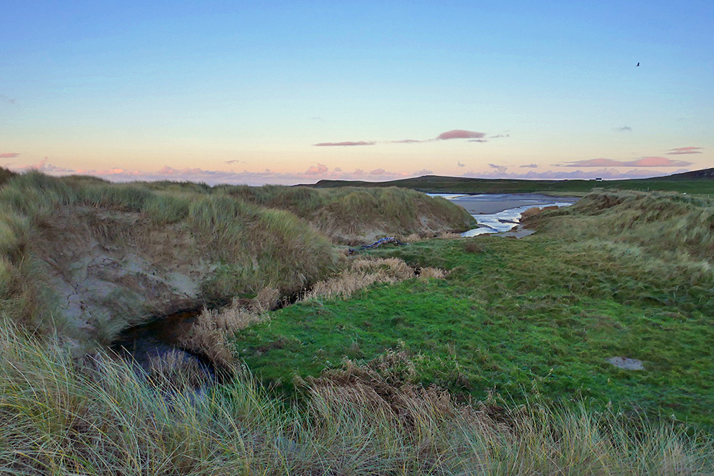 Picture of a burn (stream) curving through dunes next to a beach