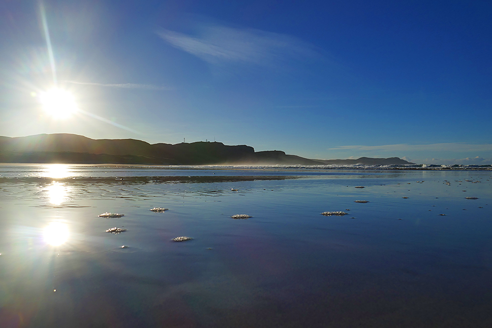 Picture of a wide sandy beach with a light layer of water under some bright low sun