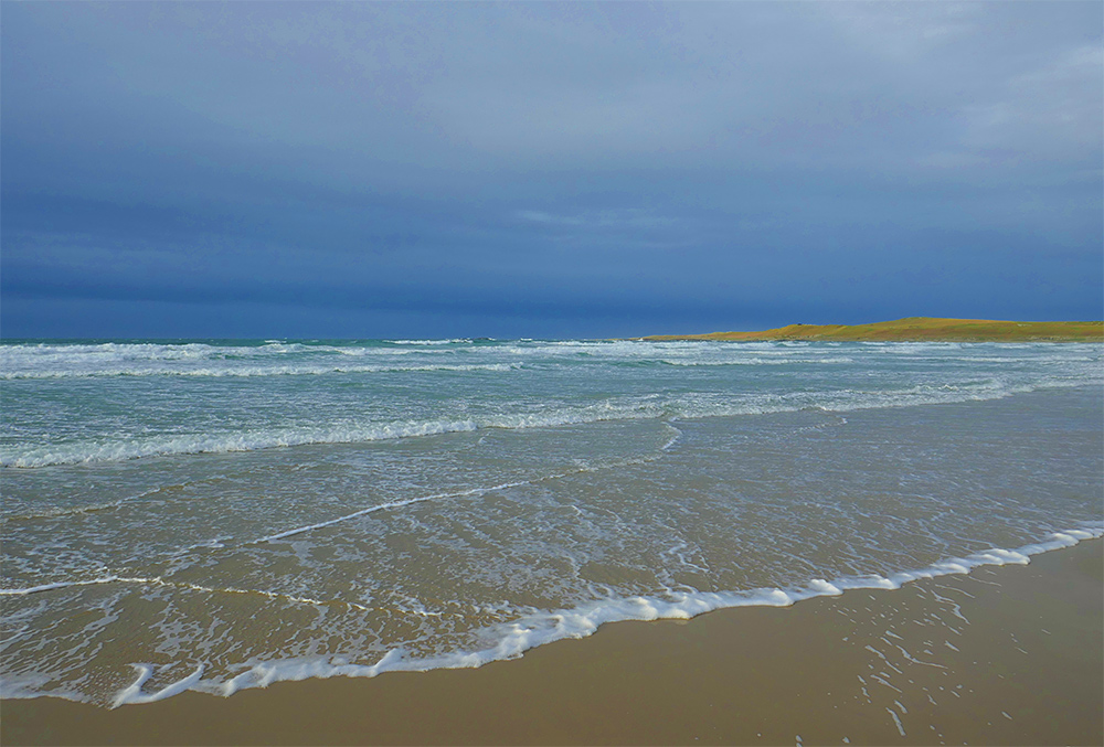 Picture of waves rolling out on a wide sandy beach in a wide bay under dark clouds