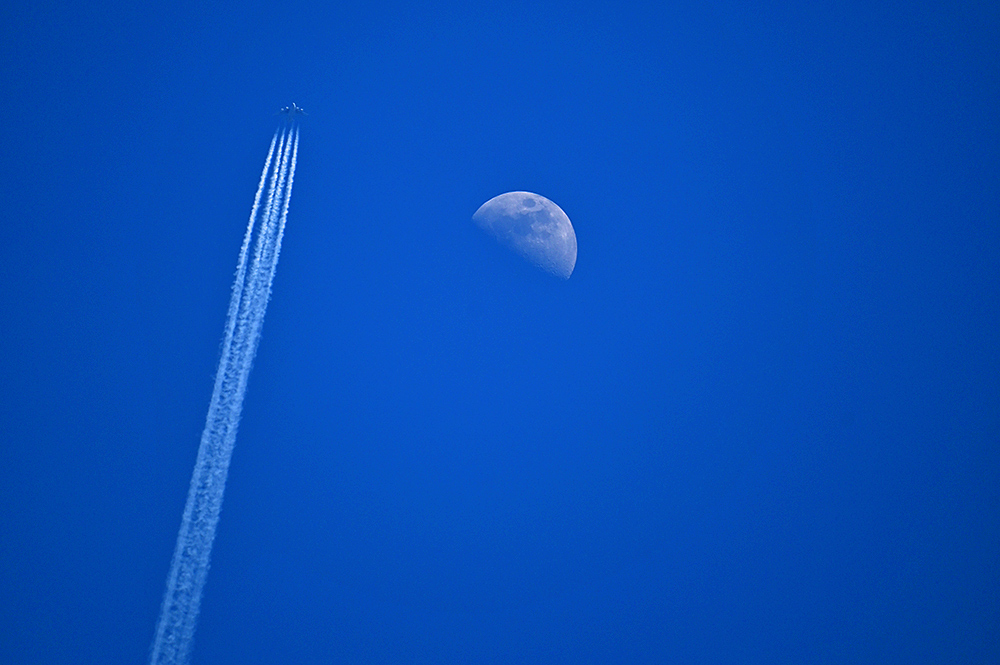 Picture of a four engine plane passing a half moon on a bright blue sky