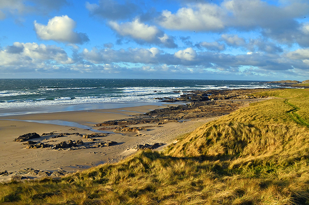 Picture of a view and over some dunes covered in dune grass down to a beach changing to a rocky shore in the distance