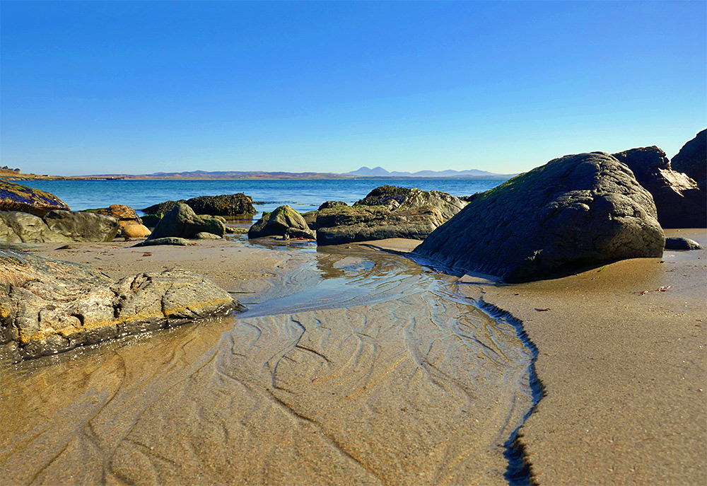 Picture of some rocks on a sandy beach, some round mountains in the distance in the background
