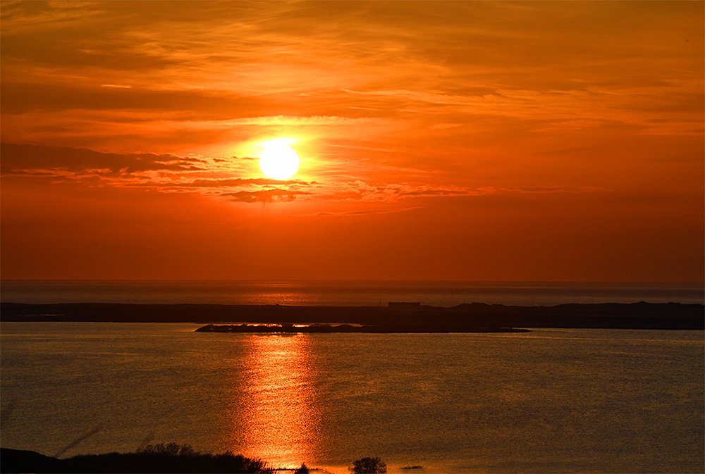Picture of a orange-red sky with the low sun visible through the clouds over sea bay next to a freshwater loch