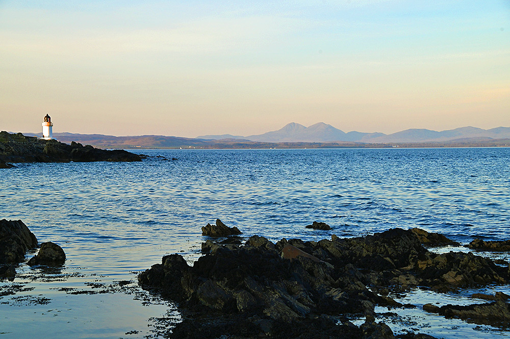 Picture of a view over a sea loch in mild evening sunshine, a small lighthouse on the shore and round mountains in the distance