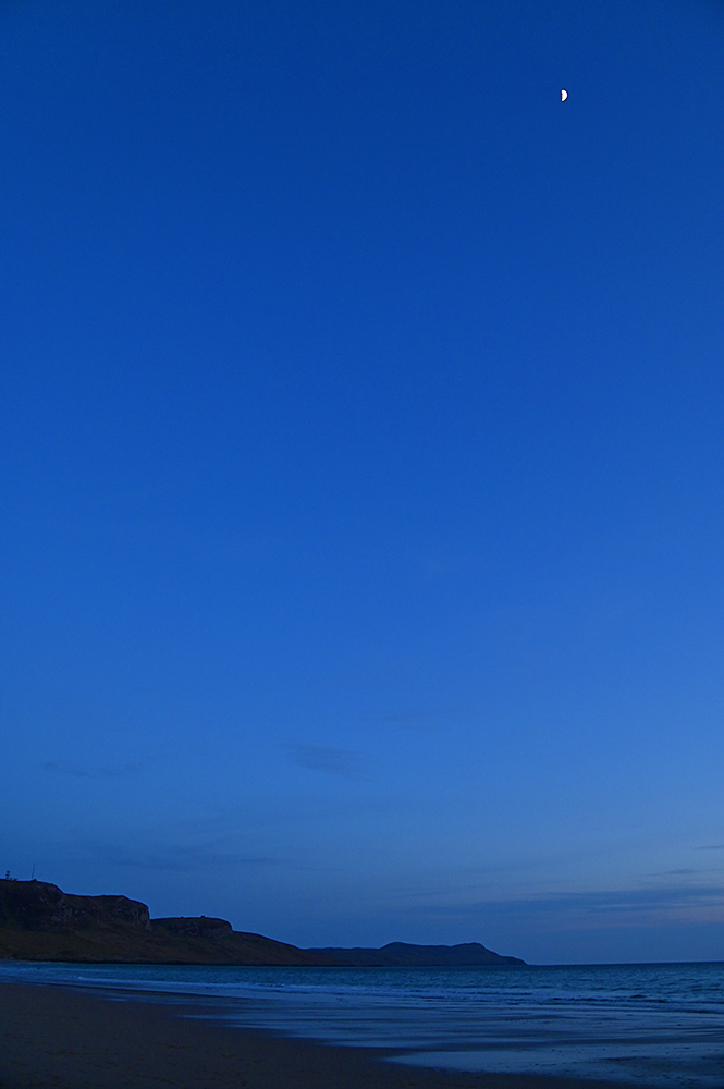 Picture of a tiny half Moon over a bay with a beach, crags and hills in the distance