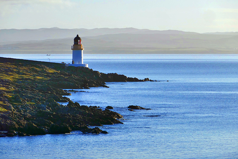 Picture of a small lighthouse on a rocky rugged shore in some mild morning sunshine, a fishing boat just visible in the distance